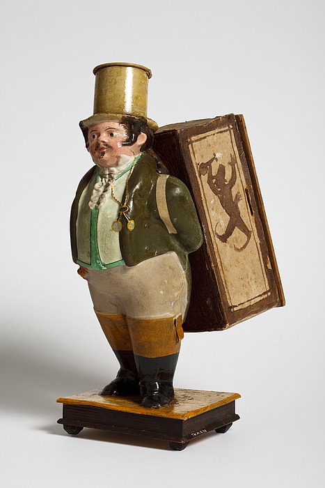 Mandlig figur med en kasse på ryggen. Indeni kassen en papirrulle hvorpå kunstnerne er tegnet gående i et optog